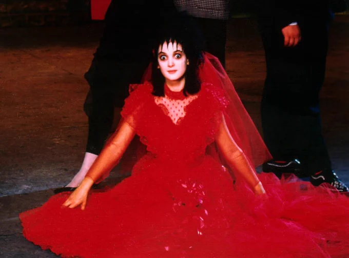 From the set of Beetlejuice. She sits on the floor in the iconic red puffy wedding dress. She's looking at the camera with wide eyes.