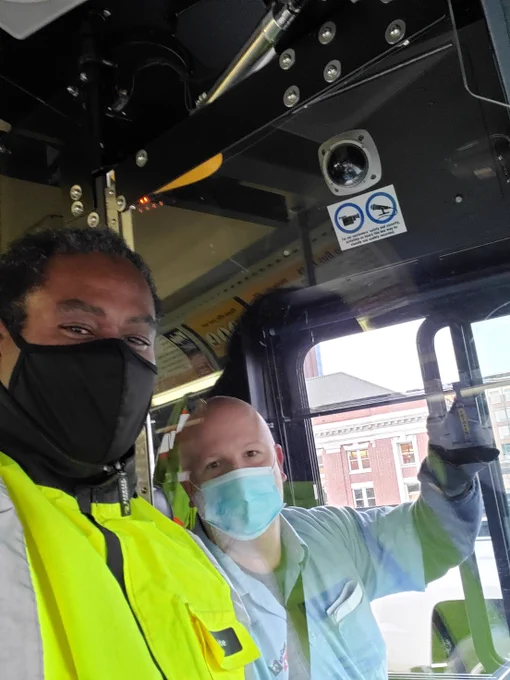 King County Metro General Manager takes a selfie with Transit Operator Patrick Getty out of South base. Terry is a tall black male wearing a black face mask and a reflective safety jacket. Over his shoulder is Patrick, sitting in the operator's seat. He's a white male wearing a blue disposable face mask and a blue Metro uniform shirt. 