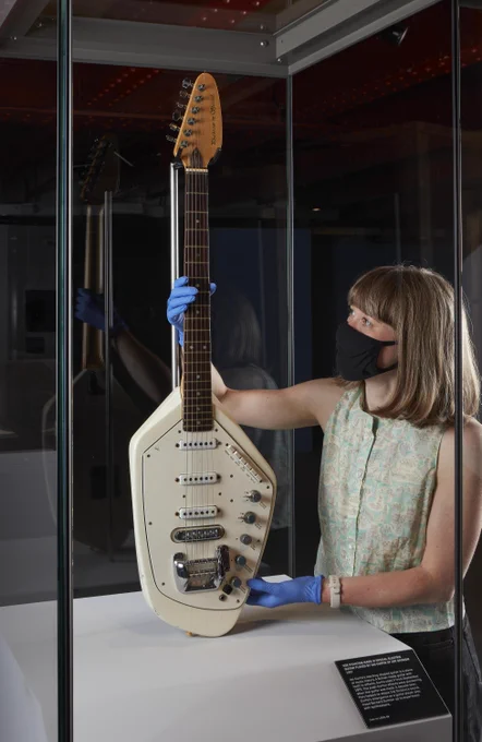 A woman places a guitar into a tall showcase. She is holding the neck upwards with blue gloved hands. The guitar has a white body and wooden neck.