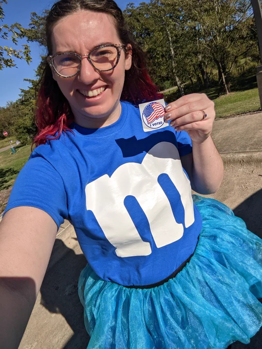 Jessica wearing a blue shirt with an m on it and blue tutu (dressed up as a blue m&m), holding an I voted sticker