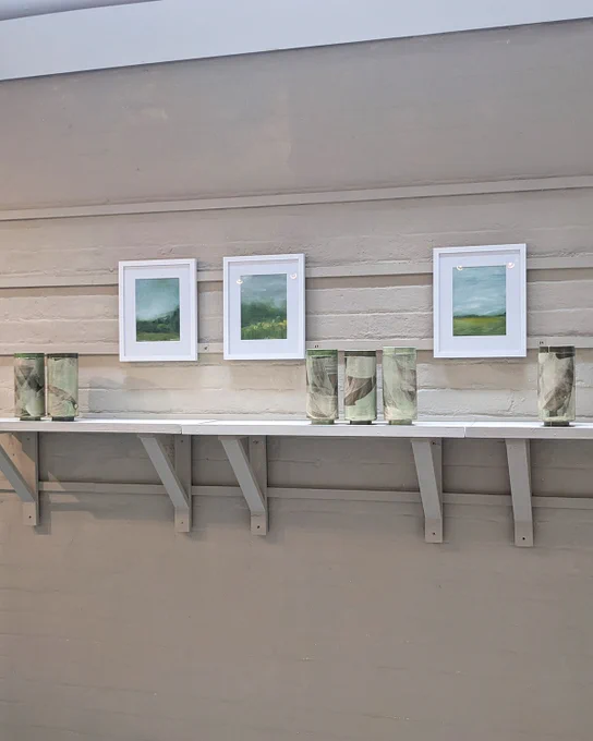 Tall, narrow, vases on a white shelf, with framed landscape prints on the wall.