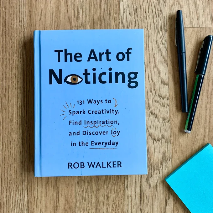 Blue book titled "The Art of Noticing. 131 Ways to spark creativity, find inspiration, and discover joy in the everyday." By Rob Walker. The book is on a wooden background with two pens and a pad of aqua sticky notes to the right.