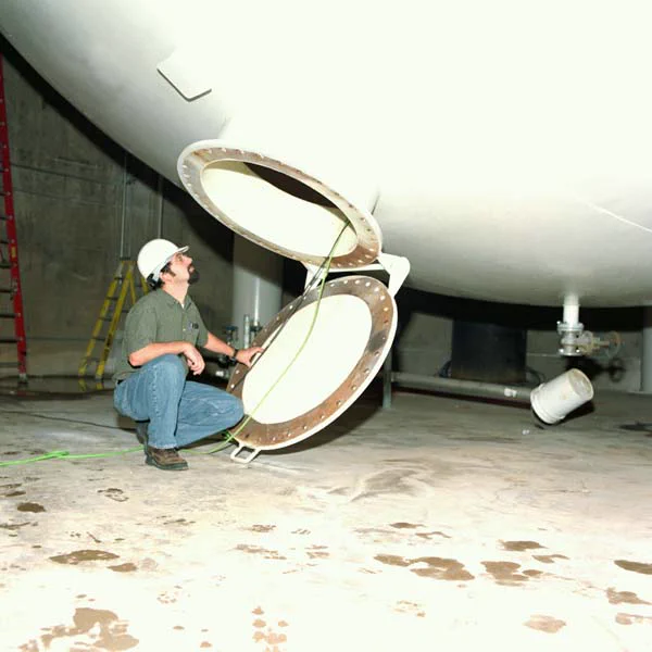 Color photo. A person in a hard hat crouched, looking up into an open hatch on a large white tank that fills about half of the frame.