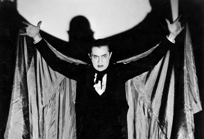 A promotional still of Lugosi as Count Dracula. He is stood in a suit looking at the camera frowning with wide eyes. His arms are out holding the cape he is wearing up. On the white background behind him we can see his shadow that resembles a bat.