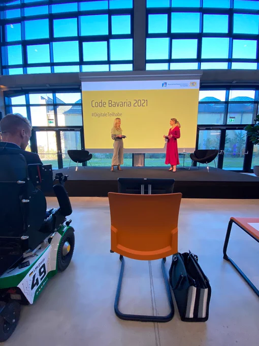 Zwei Frauen stehen auf einer Bühne und unterhalten sich.
Hinter ihnen eine grosse Leinwand in gelb auf der in schwarzer Schrift „Code Bavaria 2021“ steht. 
Vor der Bühne stehen orange Stühle, außerdem ein Mann im Rollstuhl.