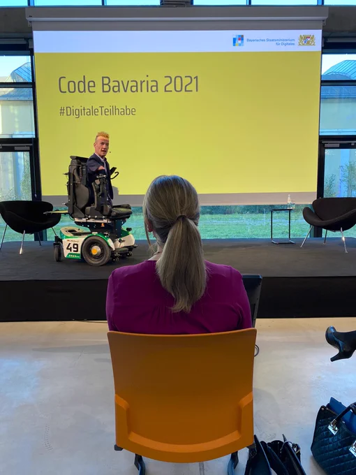 Ein Mann (Janis McDavid) hält einen Vortrag auf einer Bühne. Hinter ihm eine gelbe Leinwand mit schwarzer Schrift „Code Bavaria 2021“. Vor der Bühne sitzt eine Frau (Judith Gerlach) in einem orangen Stuhl.