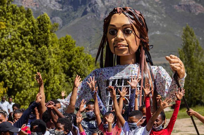 Photo of Little Amal, a 3.5-metre-tall puppet with long hair. A group of children are standing in front of her with their arms raised. There are trees and the base of a mountain or hill in the background. 