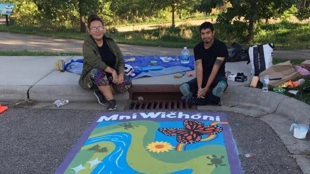 Two artists sit near their mural by a storm drain. Mural shows butterflies, flowers, and green along a river, and the words "Mní Wičóni."