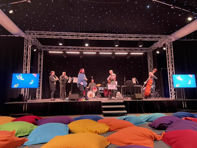 Star cloth behind the Flutter and Fly team on stage with guitar, trumpet, clarinet, double bass, percussion.. screens with butterflies on either side of the stage and brightly coloured beanbags on the floor in front of the stage. Awaiting the audience!