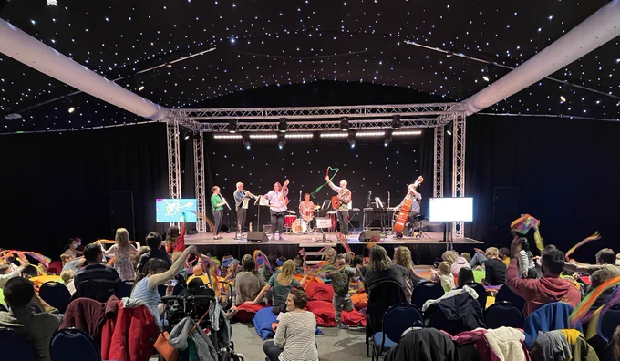 Audience of families swishing rainbow coloured ribbons in the air, led by the musicians on stage, some are playing their instruments and the others are making shapes with their ribbons. Dark black backdrop with sparkling lights on the ceiling.