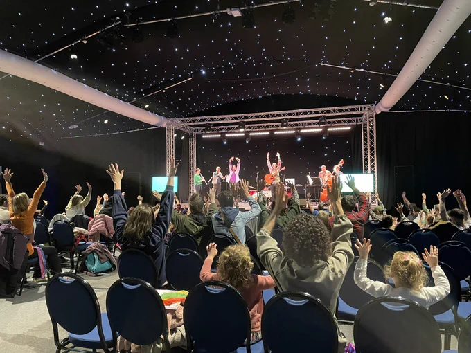 Audience of families throwing their hands up in the air! The musicians on stage are doing the same, whilst some of them play their instruments! The background is black with sparkly lights on the back wall and ceiling. The musicians include a clarinettist, trumpeter, percussionist, double bassist, guitarist & singer and a BSL interpreter.