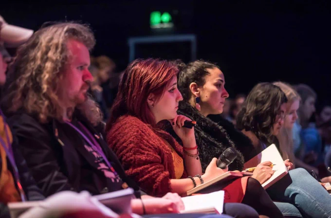 A row of attendees in an event space watch a panel. The women in the centre of the frame is listening while holding a microphone. 