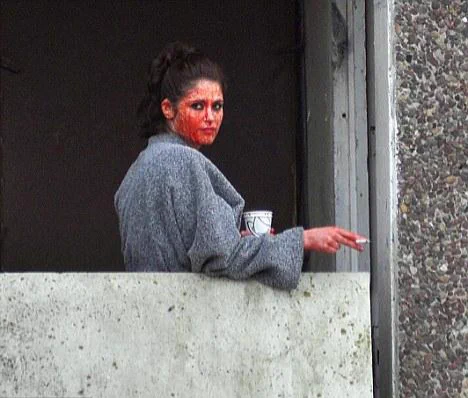 At the same balcony she is tapping the ash of her cigarette over the balcony with her back facing the camera and her face turned looking to the cameras left. She has a coffee cup in her other hand.