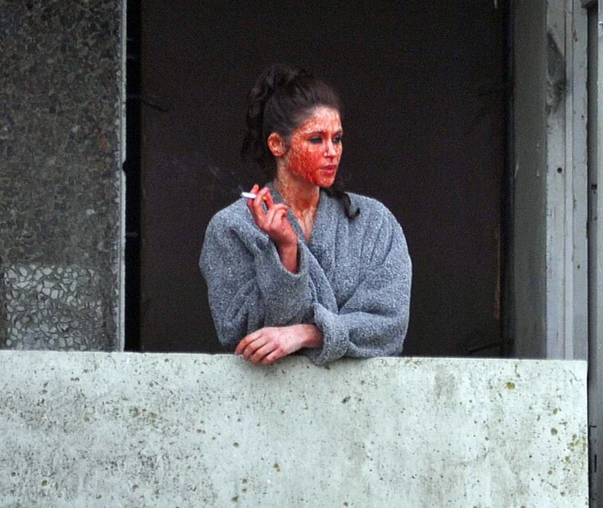 A paparazzi shot of Gemma leaning over a balcony with a cigarette in one hand. She is wearing a grey dressing gown with her face splattered with blood. She's got black eye makeup and her dark brown hair is tied back.