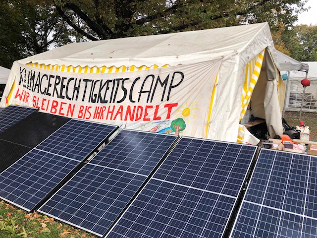 Phovovoltaikmodule vor einem Banner mit der Aufschrift "Klimagerechtigkeitscamp Wir bleiben bis ihr handelt".