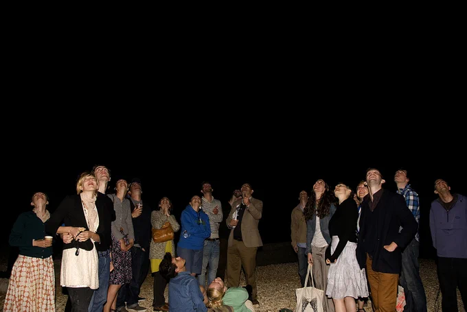 A large group of people standing in the dark on a pebble beach, illuminated only by the flash of the camera, are all looking upwards towards a spot in the dark black sky, as if watching something happen.
