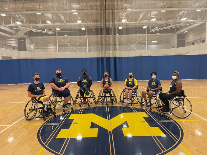 Seven wheelchair basketball players are posed at the center of a basketball court around a large yellow Block M in blue circle on the floor. The center player holds a basketball. They are all wearing  blue jerseys with yellow "M" and facemasks. 