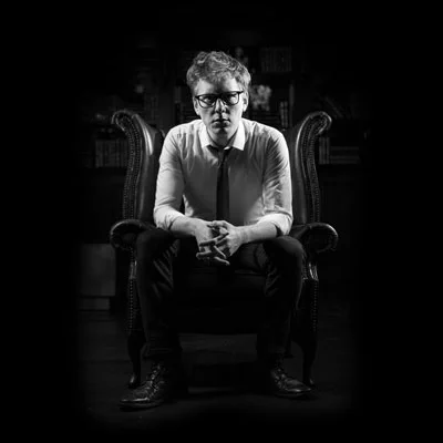 Black and white photo of a man wearing a shirt and tie, sitting in a chair, staring intently forward. 