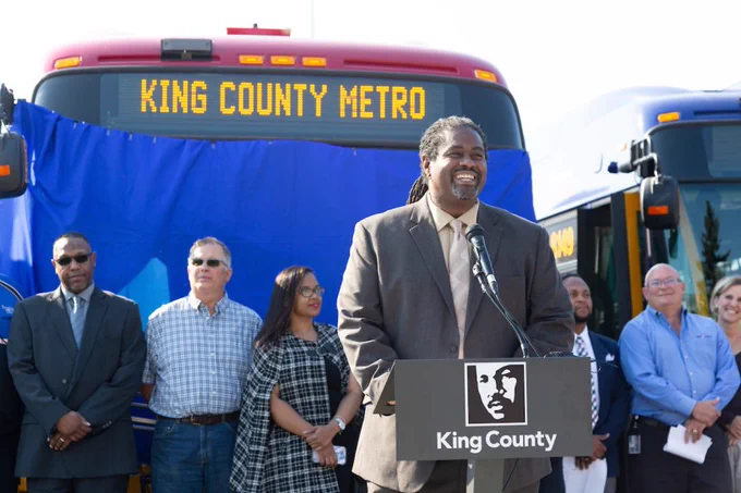 Metro General Manager Terry White stands in front of a podium that is positioned in front of a line up of people who are standing in front of a Metro bus. This 2018 file photo is from the APTA award presentation, and there is a blue banner covering the front of the bus and the award that had yet to be unveiled. Terry is a tall black male wearing a brown suit, his braids are pulled back in a low pony tail, and he has a huge smile as he addresses the crowd. 