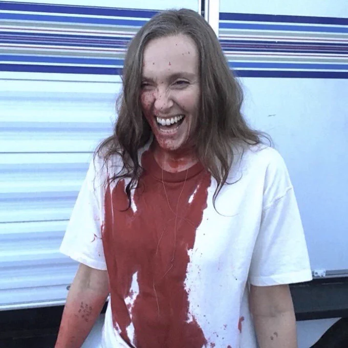 She stands in front of a trailer in a white t shirt covered in blood. She is laughing.