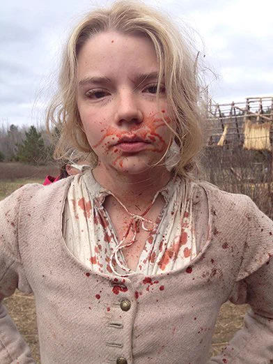 A young Anya, outside and in puritan costume, has blood splattered over her face and dress. Her hair is disheveled and she is looking directly at the camera.