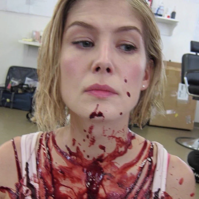 A close up photo of Rosamund in what looks like a makeup room. We can only see her head and shoulders but she is splattered in blood from her chin down.