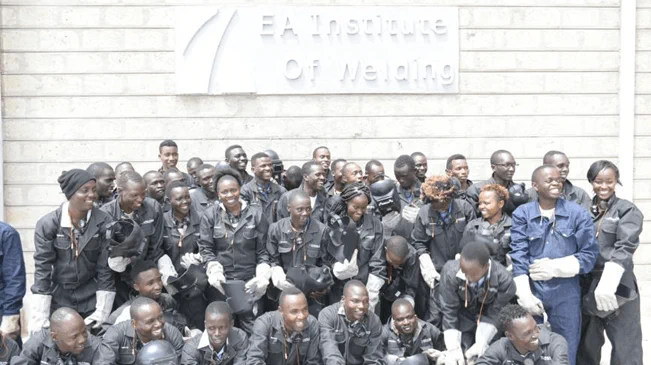 Large group of students in welding gear, engaged in dual training, posing in front of building.