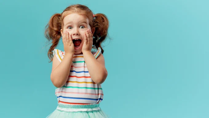 A photo of an excited child with her mouth wide open, hands pressed against her cheeks. Her hair is in pigtails and she is wearing a multicolor striped shirt.