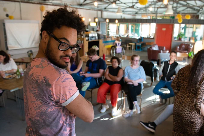 A young biracial man wearing black rimmed glasses looks back at the camera as he prepares to perform on stage for an audience.