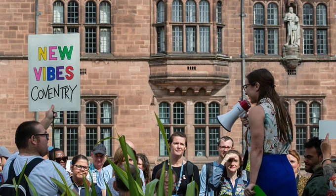 A young white woman speaks into a megaphone to a crowd outside Coventry's Council House on a sunny day.