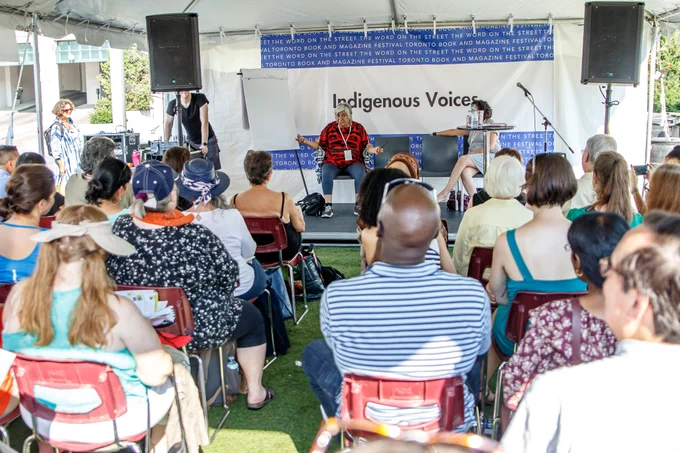 Lee Maracle speaks to a rapt audience at the 2018 Word On The Street book festival. She is wearing a black and red shirt, seated on a stage underneath a white tent, with dozens of people sitting in chairs and on the grass before her. 