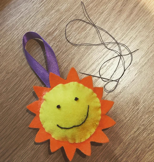 a hand made felt sun yellow with orange rays and a purple ribbon for hanging.  The sun has two eyes and a smiley face. 