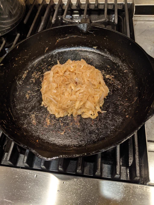 Three onions cooked down in a cast iron skillet on a stove top, much more brown than the before, but still a bit more cooking to go