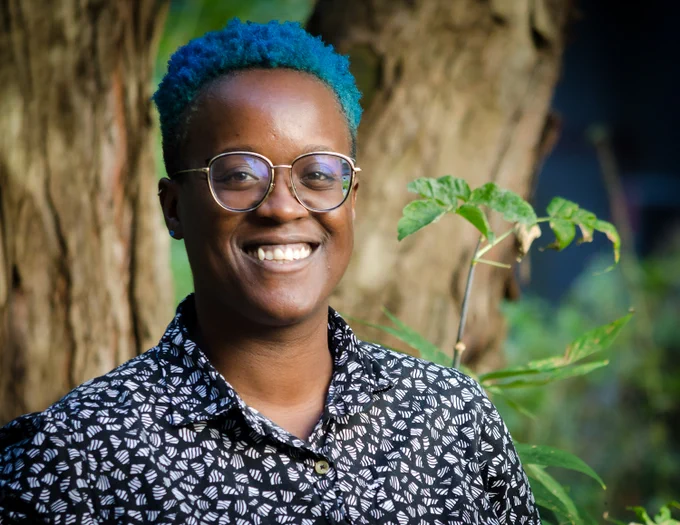 Brittany Williams. A young black woman with short teal hair stands outside and smiles at the camera. She wears a navy shirt with small white patterns. Photographed by Lysanne Larose.