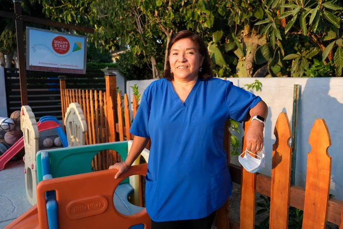 A woman in a blue shirt stands in front of children's play structures and wooden fence. 
