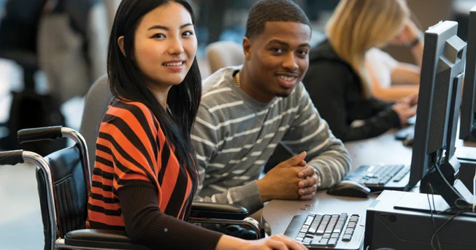 A young woman, who is a wheelchair user, sits at a computer table with a young man at work. @theABLENRC
