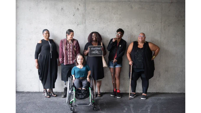 IMAGE 2 DESCRIPTION: Six disabled people of color smile and pose in front of a concrete wall. Five people stand in the back, with the Black woman in the center holding up a chalkboard sign reading "disabled and here." A South Asian person in a wheelchair sits in front. [Photo from #DisabledAndHere Project (https://affecttheverb.com/disabledandhere/) via @affecttheverb. This photo was taken by Chona Kasinger]