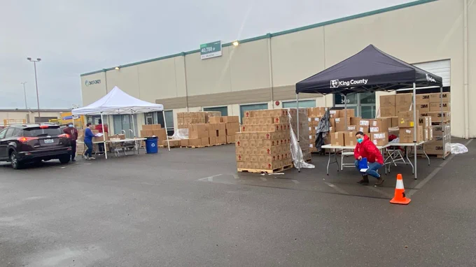 Stack of cardboard boxes stand between a white pop-up tent over folding tables and a black pop-up tent over folding tables. The black pop-up tent has the King County logo on its side. On the right, a person in red stands before cardboard boxes set up on one of the tables under the black pop-up tent. On the left, 3 people work beneath the white pop-up tent next to a dark-colored sport-utility vehicle. Behind the tents is a warehouse with beige siding and green trim.