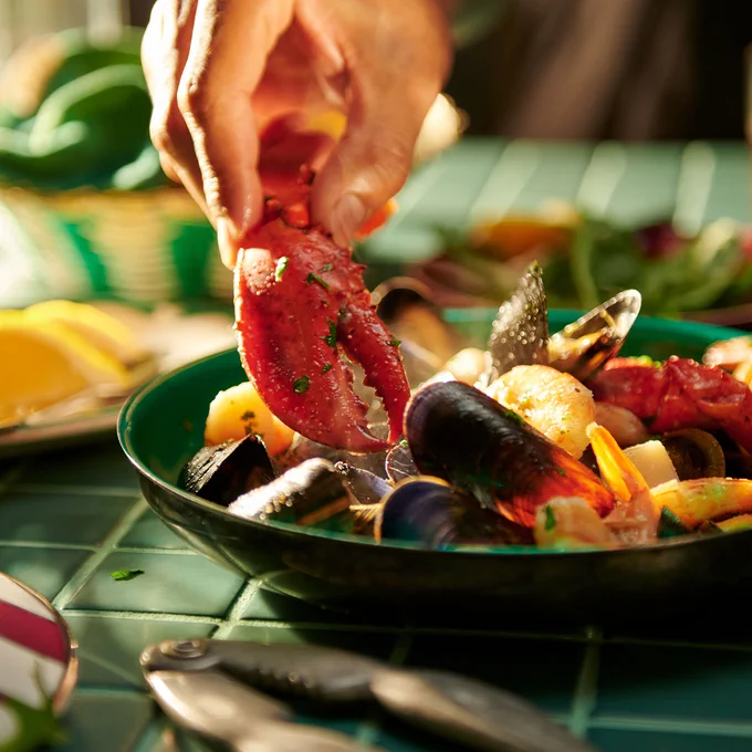 a hand grabbing a lobster claw out of a bowl of Seafood Bake. in the bowl, there are also some clams and shrimp in the bowl