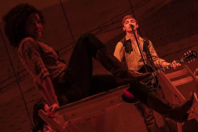 Production photo of Curly holding guitar in background, serenading a delighted-looking Laurey in foreground, all washed in red light