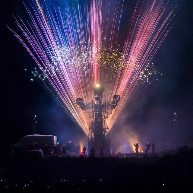 A large mechanical puppet wearing a miner's helmet with lamp stands tall above a crowd of people while fireworks light up the sky behind.