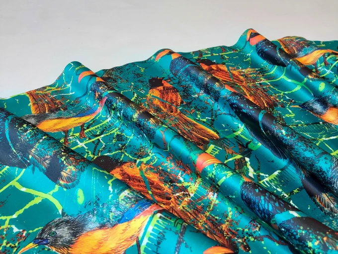 Penfia Rodrigues
ID: A close up image of a teal coloured fabric printed with bright orange, blue, green and black birds and patterns.