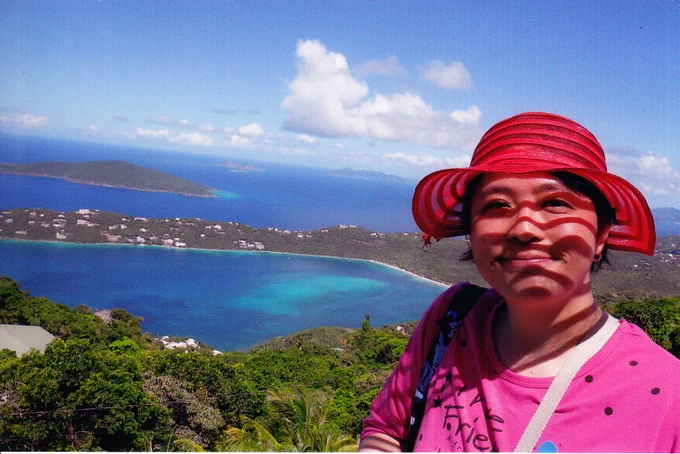 A self portrait of Rose Poon. A woman with black hair, wearing a red hat with greenery, clouds and water in the background.