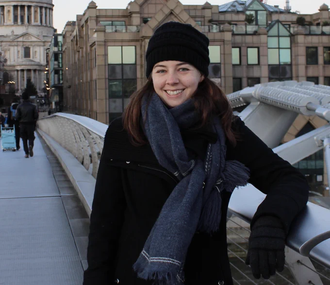 A woman with brown hair, light skin, and a black hat with a scarf smiles and poses on a bridge. 