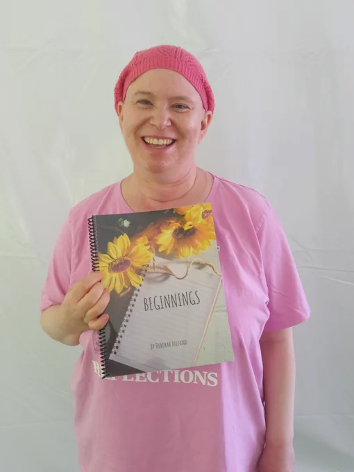 Deborah, a white person  wearing a pink shirt, and a pink knitted hat is smiling. She is holding the  book Beginnings in front of them.