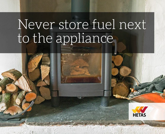 The words 'Never store fuel next to the appliance' as a banner towards the top left-hand corner. A background image of a wood burner in a brick recess with firewood stacked around it. The HETAS logo in the bottom right-hand corner.
