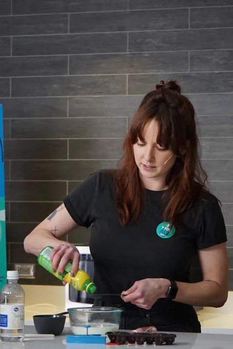 woman pouring lemon juice into a measuring cup, about to dump into bowl of milk. surrounding props: vinegar, more measuring cups, cookie cutters.