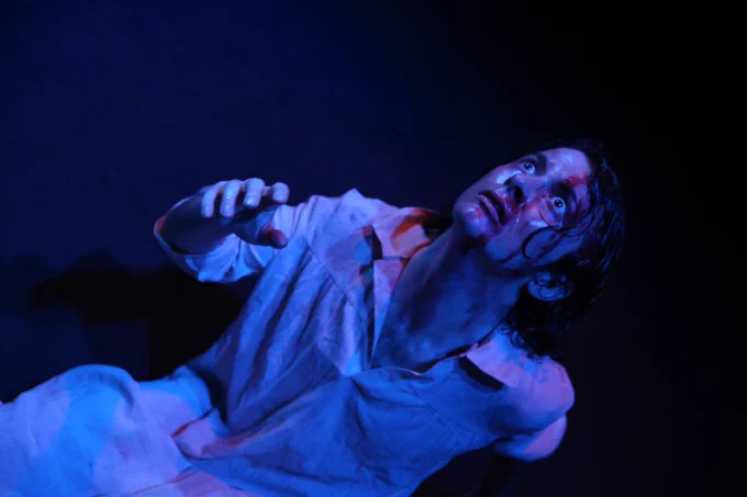 Production photo from One Man Poe. A sweaty, bruised and bloodied man in dim blue lighting, dressed in a robe, staring up at the ceiling, terrified.