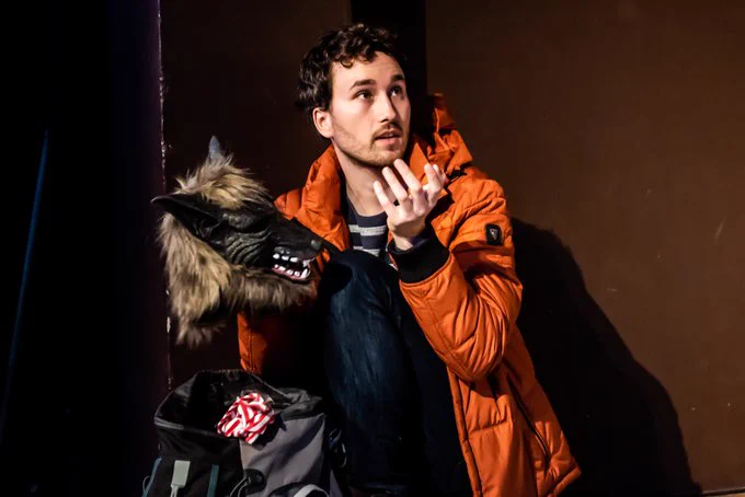 Production photo from Birdwatching. A young man is wearing a warm orange winter coat. He is crouched over a bag and holding a scary wolf mask. He is gesturing and looking at someone off camera. 