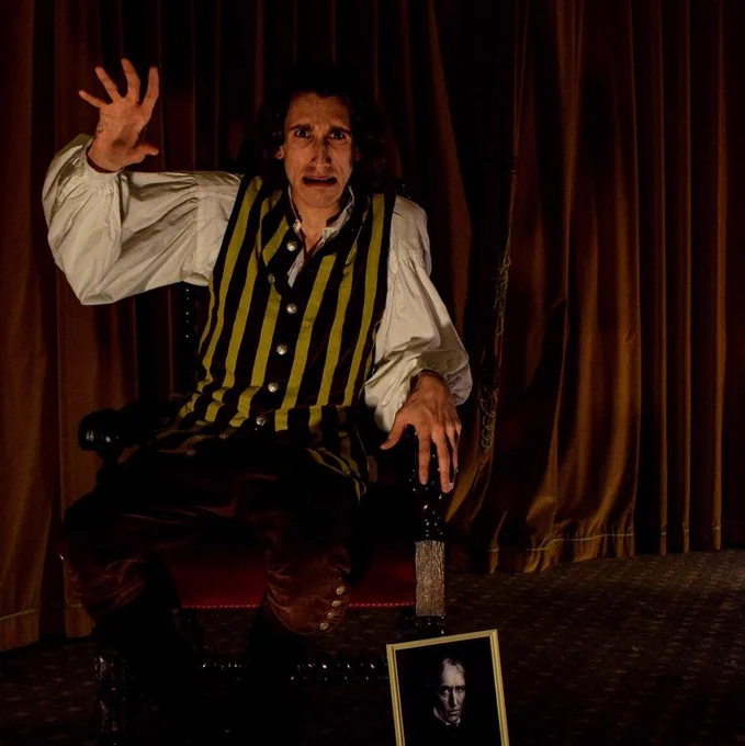 A production photo from Irving Undead. Actor James Swanton is dressed as Henry Irving, wearing a flouncy sleeved Victorian white shirt, a yellow and brown striped waistcoat, and brown breeches. He is sat in an ornately carved wooden chair with a black and white portrait of Henry Irving leaning against one of the chair legs. He has a tormented ghostly expression on his face as he wildly gestures with his arm raised and his hand wide open. The other hand clasps the arm of the chair. Behind him are dark red velvet theatre curtains.